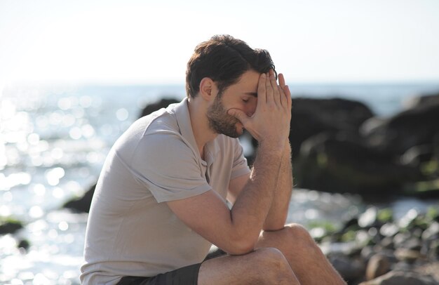 海のビーチ、顔に手のひらに座って落ち込んで悲しい若い男