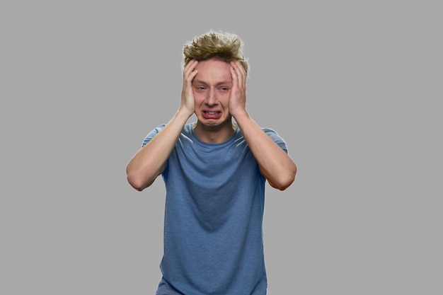 Depressed teen boy crying on gray background. Stressed emotional teen guy holding hands on head.