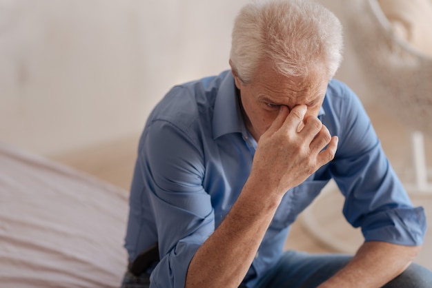 Depressed unhappy elderly man leaning forward and holding his nasal bridge while feeling unhappy