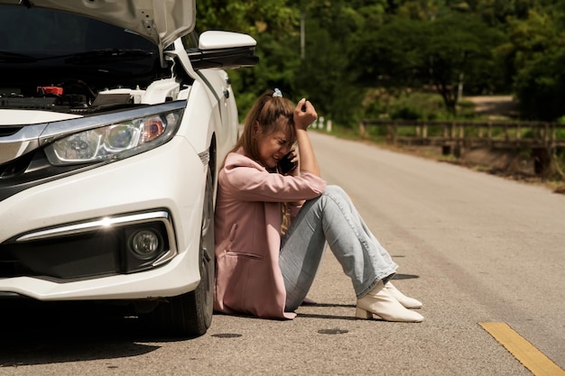 Depressed view of a young woman sitting on the road near a broken car and talking to a mechanic on the phone