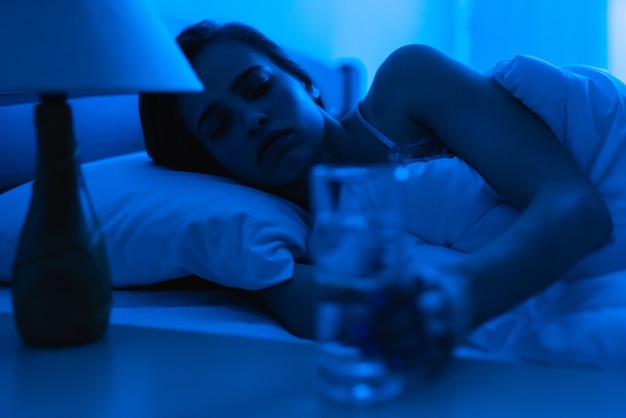 The depressed woman on the bed holding an alcohol shot. evening night time