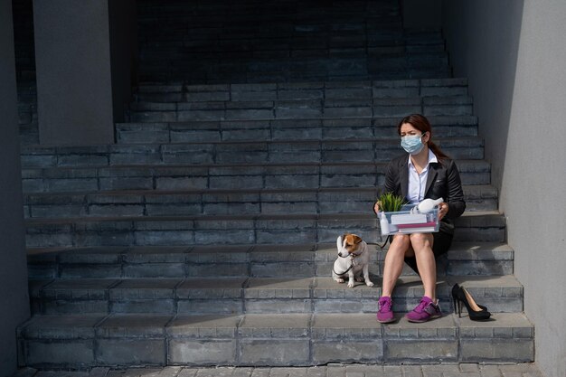 Depressed woman in medical mask is fired and is sitting on the stairs with a box of personal stuff s and a dog Female office worker in suit and sneakers outdoors Unemployment in the economic crisis