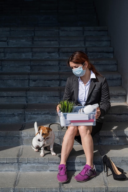 Depressed woman in medical mask is fired and is sitting on the stairs with a box of personal stuff s and a dog Female office worker in suit and sneakers outdoors Unemployment in the economic crisis