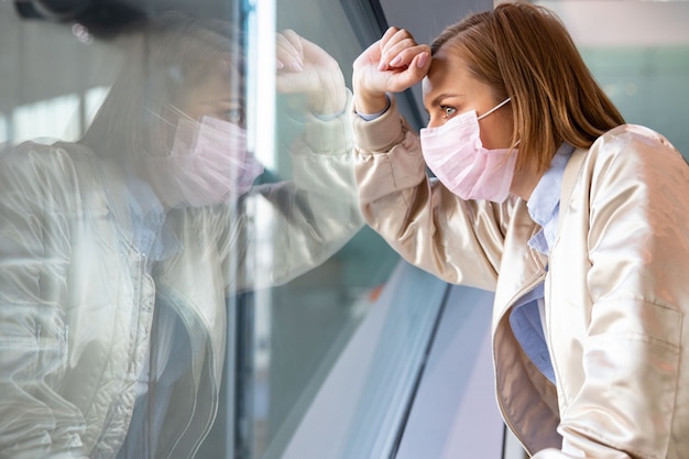Depressed woman wearing medical face mask, looking out the window at an empty city