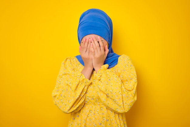 黄色の背景に大きな問題を抱えている手で顔を覆っているカジュアルなドレスを着た落ち込んだ若いアジア人女性