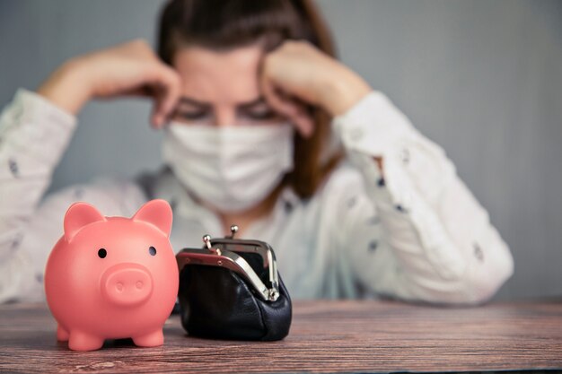 Depressed young Female business woman with pink piggy bank for savings and wearing safety mask for Covid-19