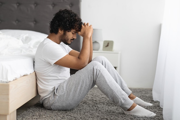 Depressed young indian man sitting by bed at home