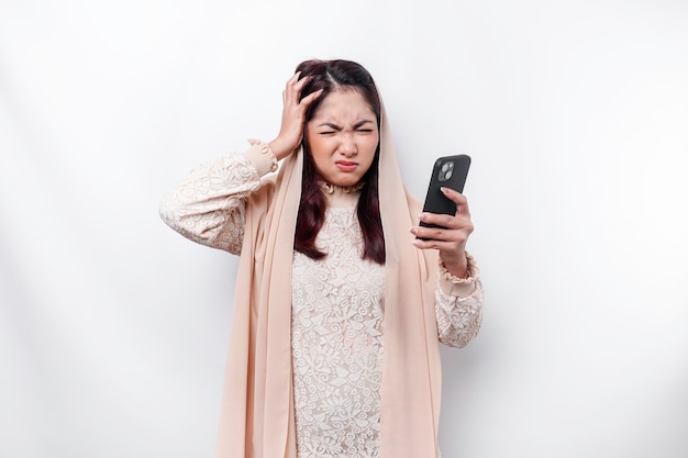 Депрессивная азиатская мусульманка в платке выглядит напряженной во время разговора по телефону на белом фоне