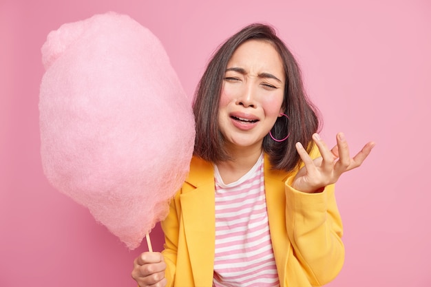 Depressief boos Aziatische vrouw heeft neerslachtige gezichtsuitdrukking houdt smakelijke suikerspin op stok voelt zich ongelukkig na ruzie met vriend draagt modieuze gele jas geïsoleerd over gele muur