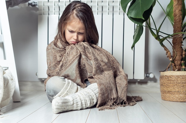 Depressief huilde een serieus klein meisje gewikkeld in een warme plaid die probeerde op te warmen en warm te blijven zitten in de buurt van de verwarmingsradiator