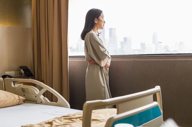 Depressieve aziatische vrouw patiënt staat is ziekenhuisafdeling kijk uit raam