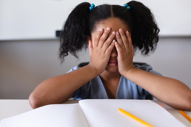 Depressieve biraciale basisschoolmeisje met handen die haar gezicht bedekken zit aan het bureau in het klaslokaal