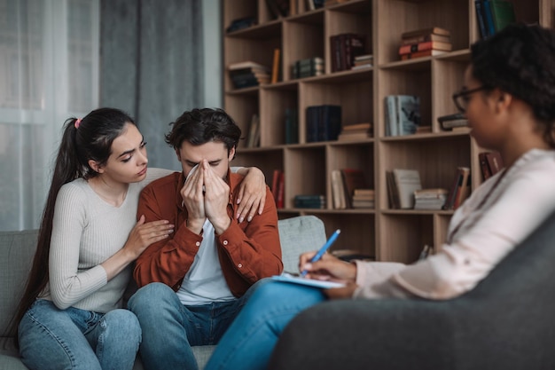 Depressieve trieste duizendjarige Europese vrouw kalmerende echtgenoot met dokter in het interieur van de kantoorkliniek