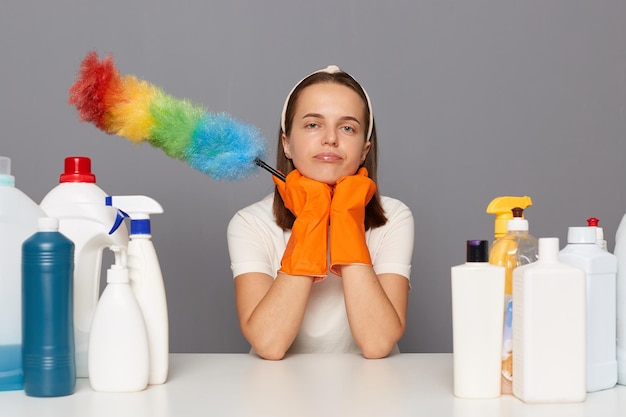 Depressieve, trieste vrouw kijkt stressvol, houdt beide handen onder de kin, draagt beschermende handschoenen en houdt een pp-stofdoek vast, omringd door veel schoonmaakproducten, voelt zich moe van de drukke dag en het schoonmaken van het huis