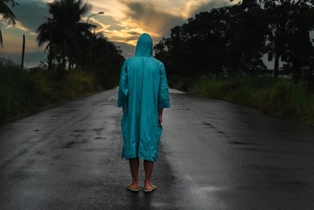비옷을 입은 외로운 남자가 일몰의 비 아래 젖은 도로에 서 있는 우울한 개념