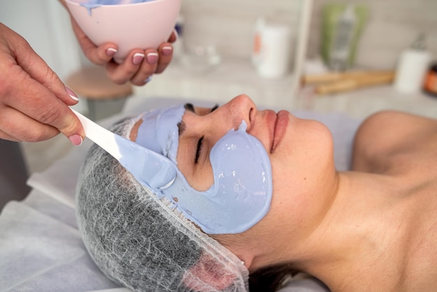 Dermatologist applying blue refreshing antiaging mask to face of young female client