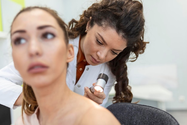 Dermatologist in latex gloves holding dermatoscope while examining attractive patient with skin disease Female dermatologist examining patient with dermascope looking for signs of skin cancer