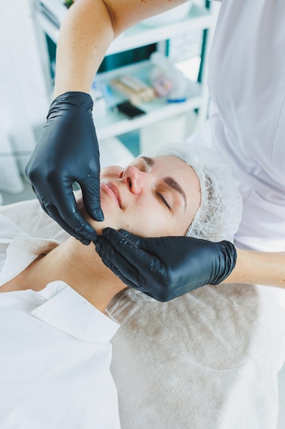 A dermatologist treats the face of a woman lying on a massage table Facial skin care A female beautician applies a mask to a womans face