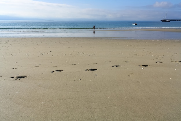 砂浜に足跡があり、海岸の隣の背景に女性がいる人けのないビーチ。