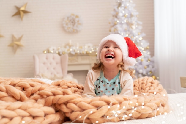 designer dekens van Merino beige! lachend kind in kerstmuts. achter het meisje op de achtergrond staat een kerstboom bij het raam. gelukkig kind liggend op het bed vreugde bij het begin van Kerstmis