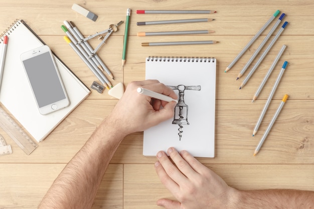 The designer draws a sketch in a notebook on a wooden table. Stationery. View from above.