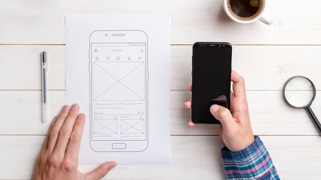 디자이너는 휴대폰에서 사용자 인터페이스와 사용자 경험을 테스트합니다. 스케치된 앱으로 휴대폰 옆에 와이어프레임이 있습니다.