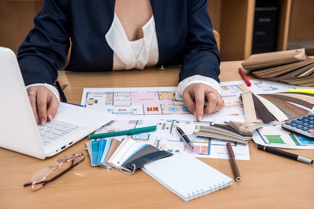 The designer works in the office with samples of colors a laptop and a plan of the building