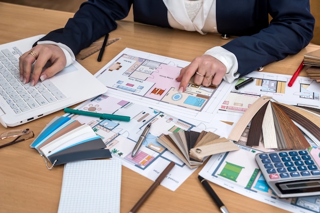 The designer works in the office with samples of colors, a laptop and a plan of the building