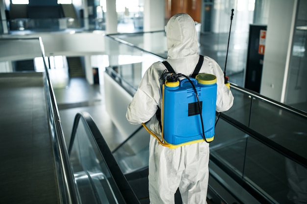 Desinfectiemedewerker die een beschermend pak draagt, ruimt professioneel een roltrap op in een leeg zakencentrum. een man uitgerust met antibacteriële kleding steriliseert winkelcentrum. covid-concept.