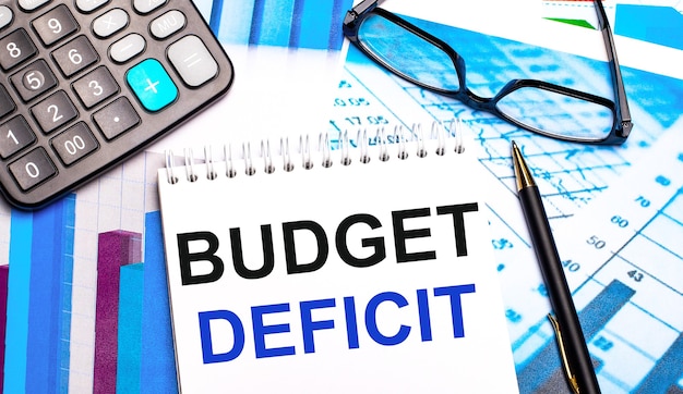 바탕 화면에는 BUDGET DEFICIT라는 텍스트가있는 컬러 테이블, 계산기, 안경, 펜 및 노트북이 있습니다.