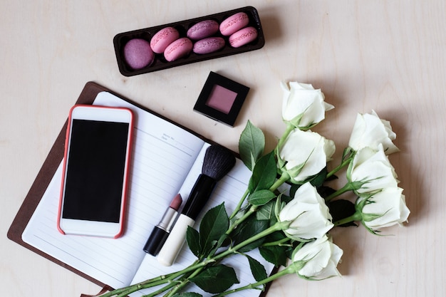 Desktop flatlay with mobile phone, notebook, macaroons, white roses, lipstick, make up brush and blush on wood