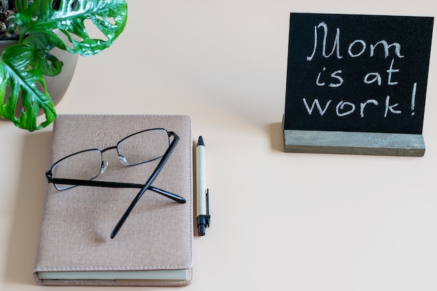 Photo desktop, home work place with time diary, pen, eyes glasses and black blank with inscription mom is at work. home remote working concept.