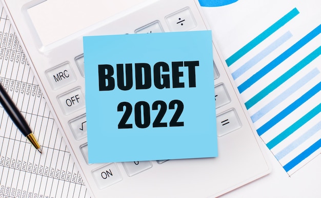 On the desktop is a white calculator with a blue sticker with the text BUDGET 2022, a pen and blue reports. Business concept