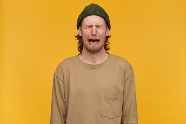 Despaired man, tearful guy with blond hair, beard and mustache. wearing green beanie and beige sweater. crying with closed eyes and feels hopeless. stand isolated over yellow wall