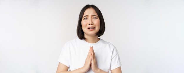 Desperate crying asian woman begging asking for help pleading and say please standing in white tshirt over white background