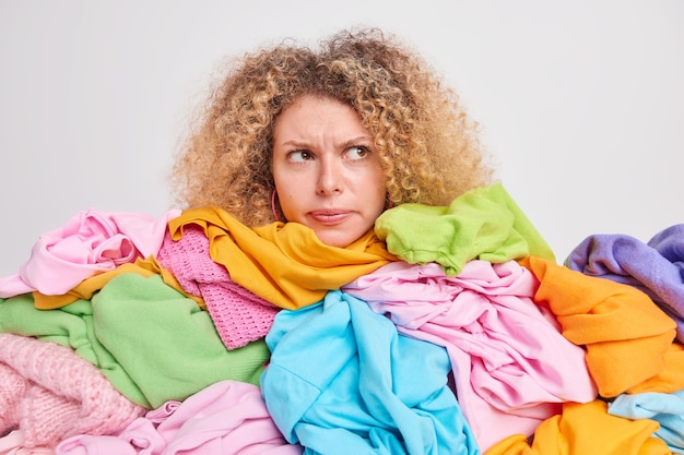 Photo desperate sad woman has curly hair being real shopaholic grabs different clothes on sale from shop overloaded with multicolored clothing focused away isolated over white wall sorts wardrobe