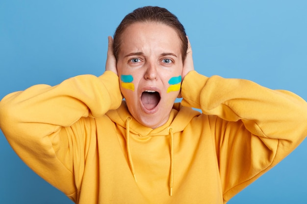 Desperate woman in yellow hoodie with Ukrainian flag on cheeks posing isolated over blue background standing and covering her ears don't want listen about war in Ukraine screaming