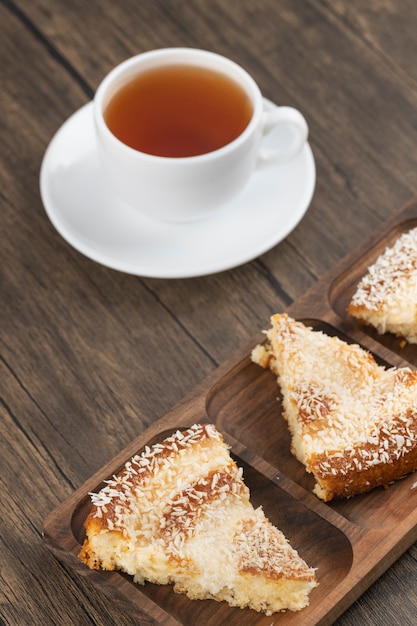 Кусочки десертного торта с кокосовой стружкой и чашка чая на деревянном столе.