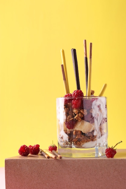 Dessert in glas met chocolade muesli, banaan en framboos, versierd met stokjes Pocky op roze en gele achtergrond, verticaal formaat, ruimte voor tekst