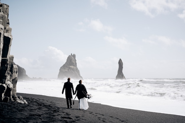 目的地のアイスランドの結婚式結婚式のカップルは近くのvikの砂浜の黒いビーチに沿って歩きます