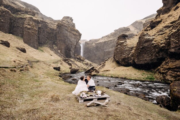 kvernufoss滝の近くの目的地のアイスランドの結婚式結婚式のカップルが川のほとりに座っています
