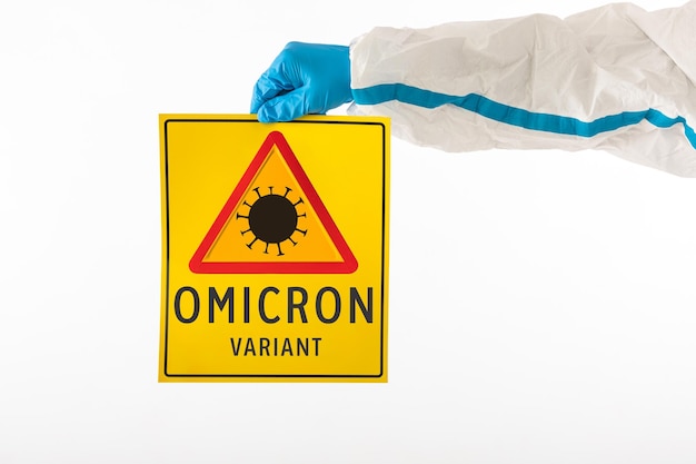 Detail of an arm of a medical nurse wearing a PPE and latex gloves with a yellow sign with a COVID-19 danger symbol that reads: 'Omicron variant'. Coronavirus, pandemic and health concept.