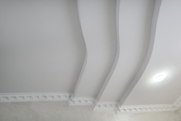 Foto particolare del soffitto ad angolo con intricate modanature a corona soffitto sospeso e costruzione in cartongesso in una stanza vuota in un appartamento o in una casa soffitto teso bianco e di forma complessa