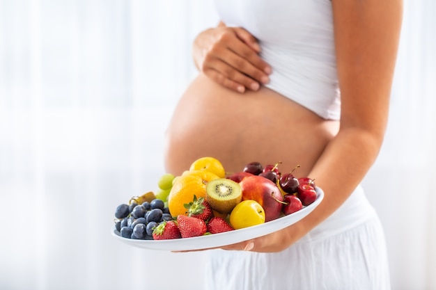 Particolare del piatto di frutta tenuto da una donna incinta con uno sfondo bianco.