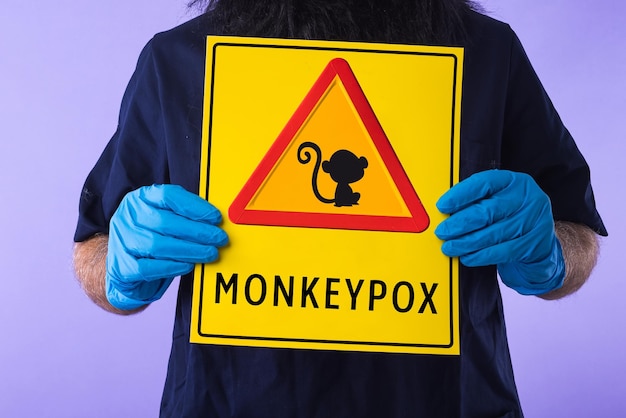 Detail of the hands of a person dressed in a doctor39s outfit holding a sign with the danger sign and a monkey that says MONKEYPOX on a purple background Virus epidemic Nigeria and smallpox