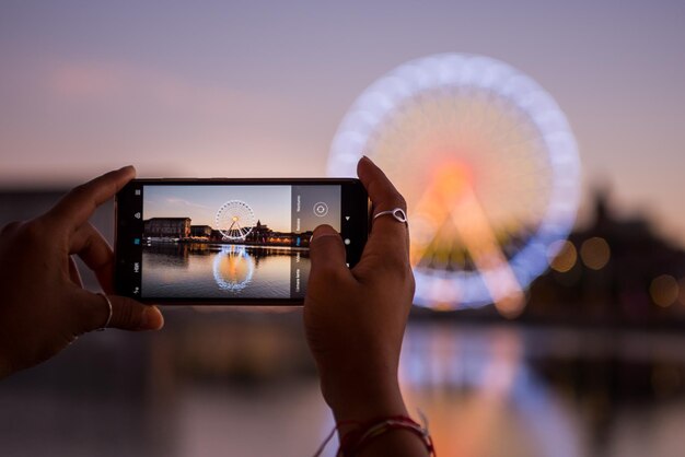 Foto dettaglio delle mani che scattano una foto della ruota panoramica con un telefono cellulare