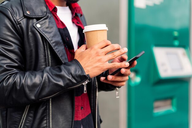 Detail of the hands of an unrecognizable modern young man consulting his phone at the bus stop with a take away coffee in his hand, concept of technology and urban lifestyle