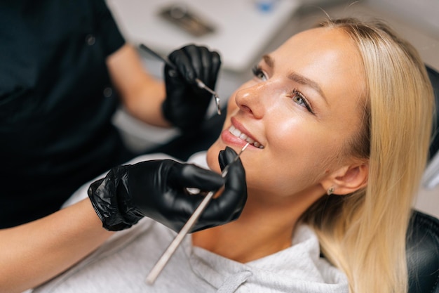 치과 클리닉에서 치료를 받고 있는 행복한 금발 여성 환자의 세부 헤드 클로즈업 장갑을 입은 인식할 수 없는 치과의사의 손이 환자의 치아를 검사하고 있습니다.