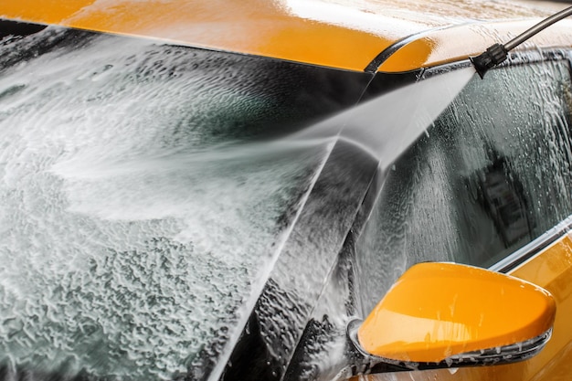 Detail op de donkergele voorruit van een auto en de zijspiegel die wordt gewassen met waterstraalspray, zeepschuim.