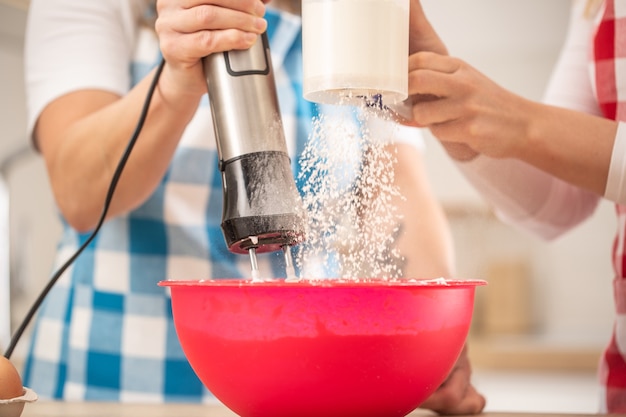 Detail of two females putting flour into a red bowl, mixing them.